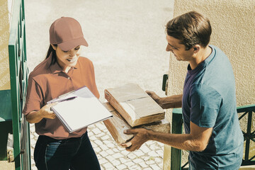 Cheerful post office worker delivering carton boxes to client, smiling and holding clipboard for signing. Attractive man receiving order at home. Express delivery service and online shopping concept