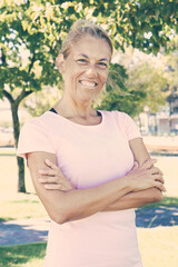 Cheerful fair haired sporty mature woman with crossed arms standing outside, looking at camera and smiling. Vertical shot. Active lifestyle concept