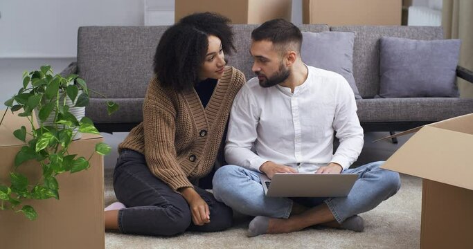 Concentrated caucasian man and happy afro american woman sitting on living room floor near cardboard boxes of parcels looking at laptop screen using computer for online shopping looking for apartment