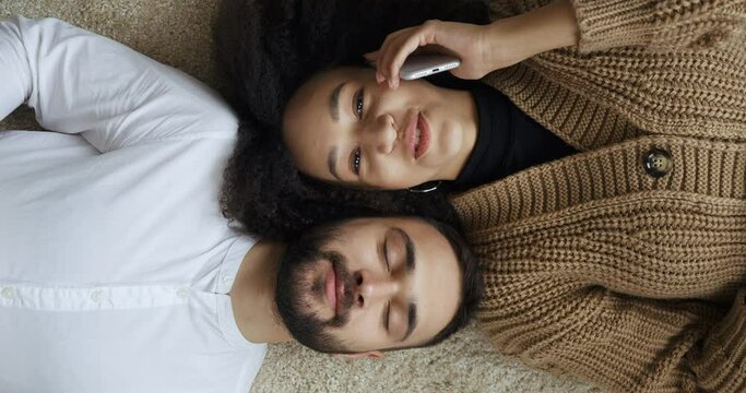 Top view afro american woman and caucasian man lying on floor, multiethnic couple resting at home girl talking on phone to mobile gadget device guy making smile repeats her words says simultaneously