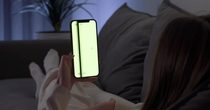 Young woman rests on the sofa and uses smartphone, scrolls through the images and swipe across the green screen, female on self-isolation, chromakey.