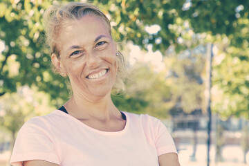 Happy positive sporty mature woman with folded arms standing outside, looking at camera and smiling. Vertical shot. Active lifestyle concept