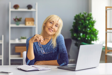 Positive middle aged blonde woman worker sitting in office at desk with laptop and notes and looking at camera during working day. Freelance, online distant working, job, occupation, manager concept