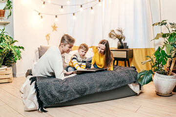 Happy family mother, father and boy at home in bed read the book. Green houseplants around, sustainable design. Yellow and gray colors 2021 year.