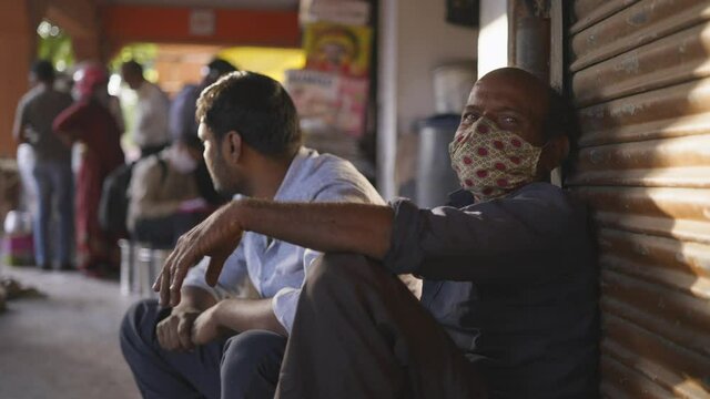 indian man sitting outside a closed city local shop wearing hand made face mask staring at lens during the hard times of covid 19