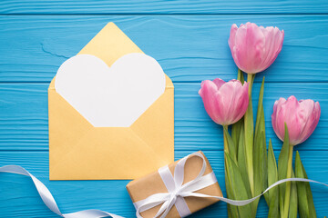 Happy valentine's day concept. Flat lay top view photo of beautiful bouquet of tulips wrapped giftbox and golden open envelope with paper card in shape of heart with mock up space on bright color desk