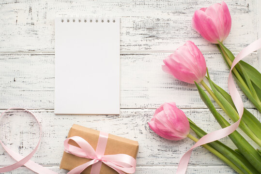 Flat lay photo picture of delicate tender tulips wrapped in craft paper giftbox surprise and notebook with empty blank clear paper page for mock up isolated vintage wooden table