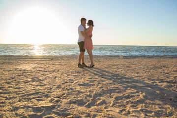 Beautiful young couple kissing on the beach at dawn. A guy in a white t-shirt hugging with a girl in a short pink dress on an empty beach near a calm sea in the early morning in summer.