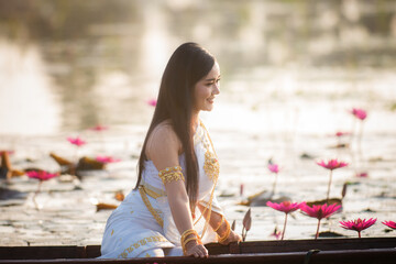 Beautiful girl in White Thai dress Sitting on a wooden boat in the red lotus pond.Thai girl in retro Thai dress,Beautiful Thai girl in traditional dress costume
