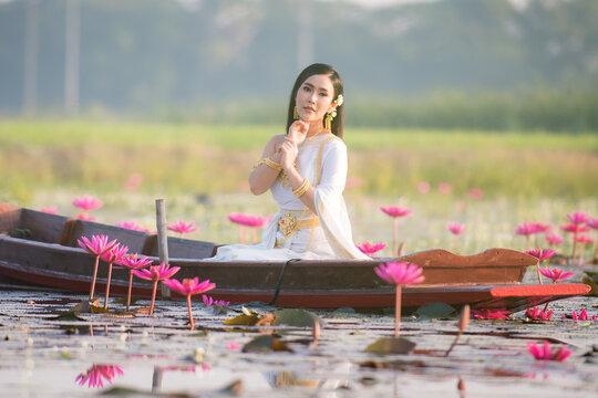 Beautiful girl in White Thai dress Sitting on a wooden boat in the red lotus pond.Thai girl in retro Thai dress,Beautiful Thai girl in traditional dress costume