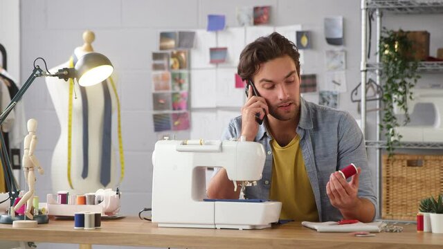 Camera pans across fashion studio as male clothes designer at sewing machine talks on mobile phone - shot in slow motion