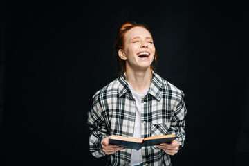 Laughing young woman college student with closed eyes holding opened book on black isolated background. Pretty redhead lady model emotionally showing facial expressions in studio, copyspace.