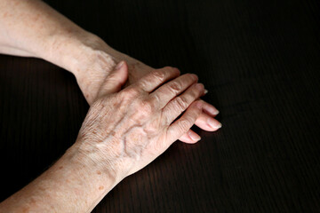 Wrinkled hands of elderly woman on a dark wooden table close up. Concept of old age, arthritis,...