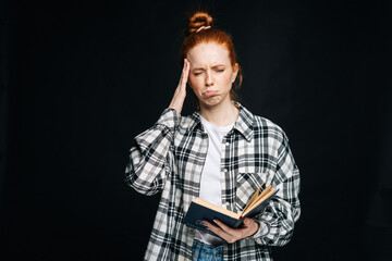 Naklejka premium Crying upset young woman college student with headache holding book and touching temples on isolated black background. Pretty redhead lady model emotionally showing facial expressions in studio