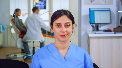 Obraz na płótnie Canvas Dental assistant looking at camera while doctor examining patient in background. Professional stomatologist nurse smiling on webcam sitting on chair in waiting room of stomatological clinic.