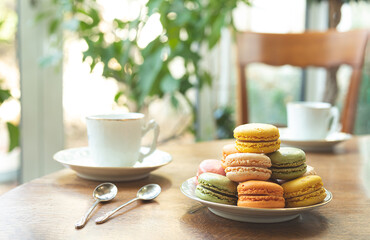 Multicolored macarons or macaroons, traditional French dessert on a plate on a round wooden table with white cups, tasty breakfast