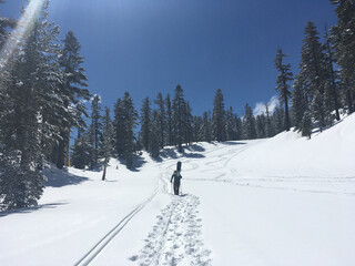 A snowboard hikes up a mountain with poles and board on his back, leaving boot tracks in the snow