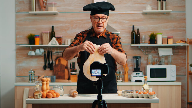 Baker using wooden rolling pin for dough in front video camera recording new cooking episode. Old blogger chef influencer using internet technology communicating on social media with digital equipment