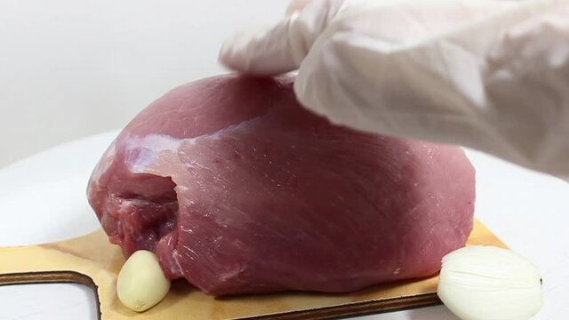 A gloved hand presses on a raw piece of pork that is lying on a garlic chopping board. Selective shot.