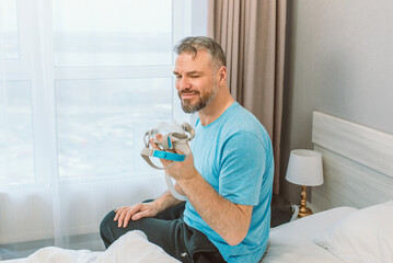 Mature happy man with chronic breathing issues considers using sitting on the bed in bedroom with...