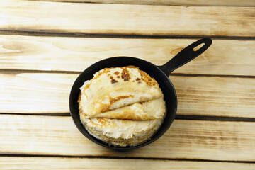 Thin pancakes in a rustic black frying pan on a wooden background.