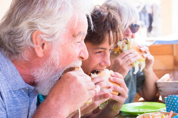 DIfferent ages group of people family eat together hamburger - grandfathers and grandson enjoy food from the table - junk and no healthy sandwich caucasian men and woman lunch concept