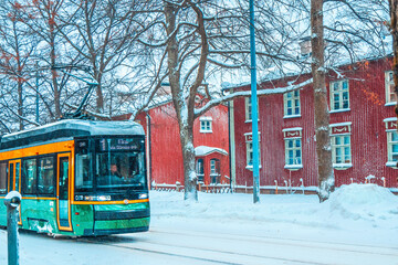 Fototapeta na wymiar Modern green tram on a snowy winter street. Old colorful building facades at street covered in snow in winter city. Red houses. Transportation concept. Contrast of old and new. Snowfall Road Helsinki.