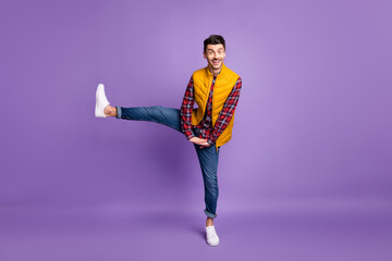 Full length photo of cute cheerful young guy dressed yellow vest dancing rising leg isolated purple...