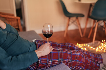 Man in check pyjamas holding a glass of red wine, cosy living room with candid light