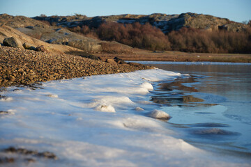 A frozen lake with ice and gravel on the shore in the foreground and scaurs in the background