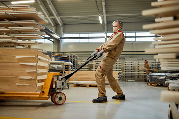 Manual worker with face mask pulling pallet jack with wood planks at carpentry workshop.