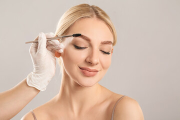 Young woman undergoing eyebrow correction procedure on grey background