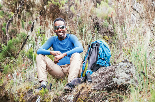 Portrait of a cheerfully smiling African-American Ethnicity young man in sunglasses sitting with a backpack, resting in the forest and looking at the camera. Active people and traveling concept.