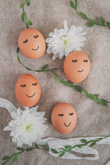 Fototapeta na wymiar 4 easter eggs with painted faces and smiles on a light beige background with white flowers and a green ribbon from plants, spring and easter concept flat lay, vertical photo 