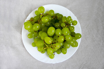 Grona of white seedless grapes in a white plate on a beige linen tablecloth top view . raw fruit on a plate