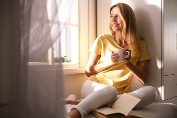 Beautiful young woman with cup of drink near window at home