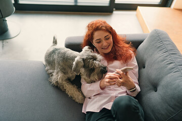Portrait of smiling young woman relaxing on sofa, using mobile phone and playing with a dog