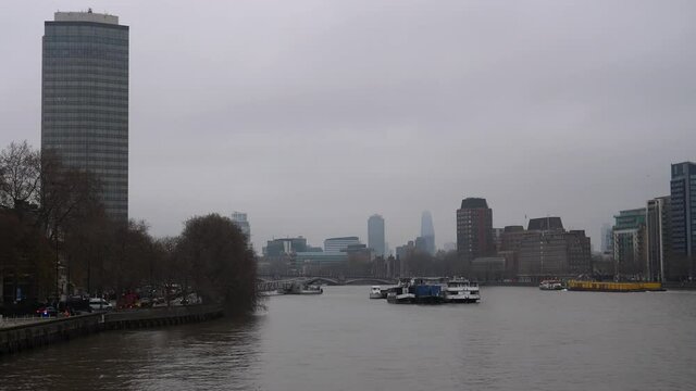 View of city from Vauxhall Bridge over River Thames in London. Motor boat is towing large platform with heavy load, black unmarked car is flashing blue lights rushing on riverside road on the left.