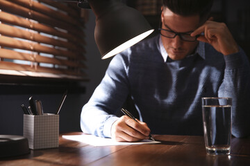 Man writing letter at wooden table in dark room, focus on hand