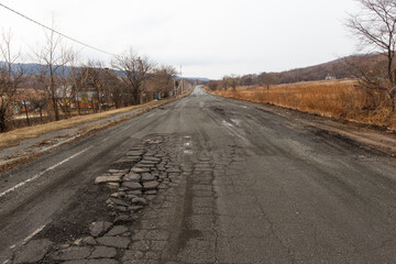 Bad Russian asphalt road. The asphalt road is full of holes and cracks among dried autumn trees.