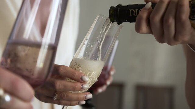 On the holiday, a young white girl holds a transparent clean glass decorated with beads and an elegant pattern, and another person pours champagne with foaming bubbles.