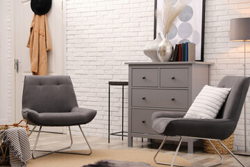 Stylish room interior with grey chest of drawers and chairs near white brick wall