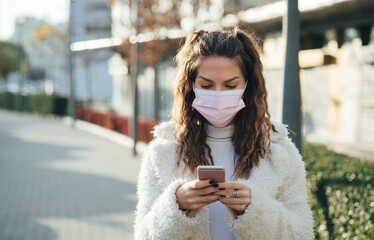 Serious young woman with pink mask checking her phone on the street