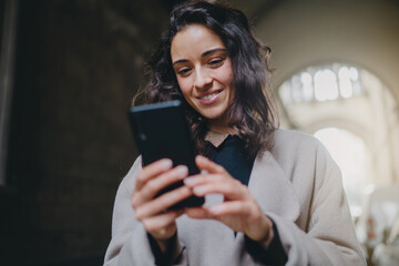 Low angle shot of young good-looking smiling woman wearing mask and light-colored coat. Good vibes while walking city streets on sunny day and using smartphone to chat. Modern technology advantages