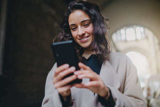 Low Angle Shot Of Young Good-looking Smiling Woman Wearing Mask And Light-colored Coat. Good Vibes While Walking City Streets On Sunny Day And Using Smartphone To Chat. Modern Technology Advantages