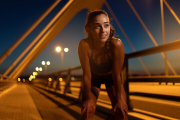 Young female athlete stopped to regain breath after running over bridge at twilight. Sportswoman puts her hands on her knees after morning jogging routine to recover. Sports and health concept
