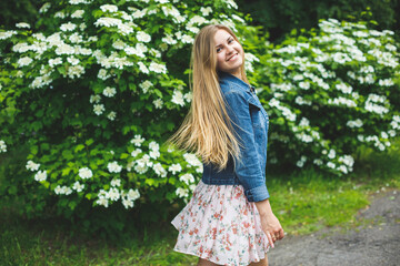 Fototapeta na wymiar A young woman of European appearance with long blond hair, dressed in a short dress, stands against the background of white flowering bushes. Sunny spring day. Natural female beauty