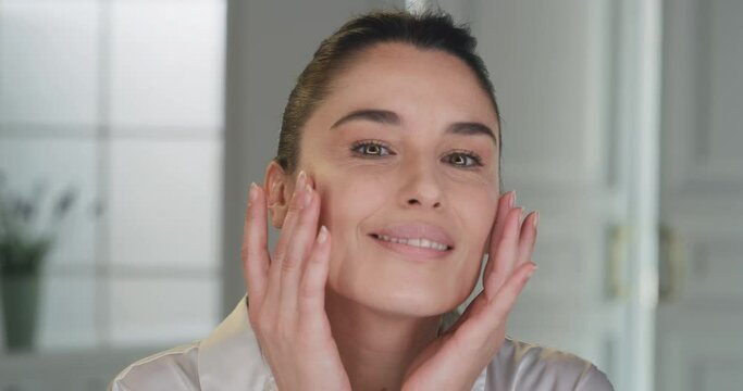 Beauty shot of mature woman with perfect face skin after applying moisturizing cream pampering it gently with fingers to absorb better on golden background. Concept of skincare, cosmetics, healthcare