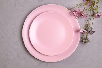 Close-up flat lay of empty pink plate and sprig of pink wildflowers on rough gray concrete or cement background. Spring concept. Selective focus.