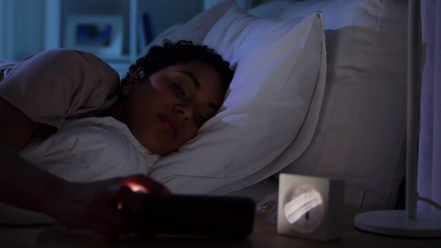 technology, internet, communication and people concept - young african american woman sleeping in bed awaking because of smartphone at home at night